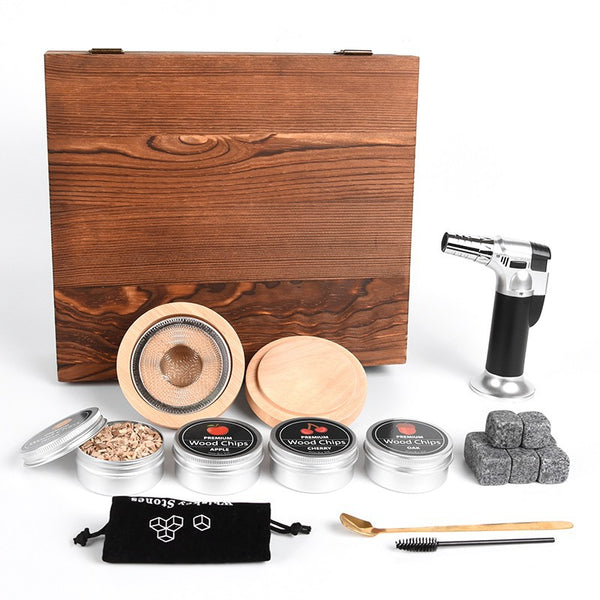 Cocktail smoker set laid out: wooden box, large-mesh smoker top and lid, torch, four wood-chip tins, chilling stones, pouch, spoon and brush.